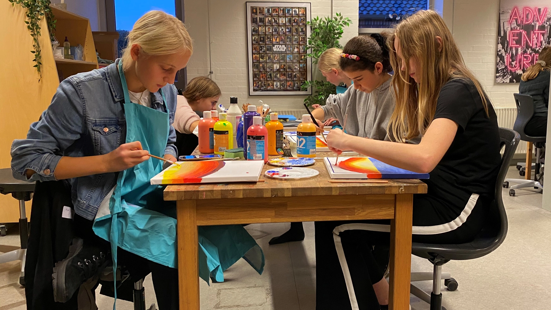 Teenagers painting on canvases while seated on a rectangular table